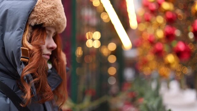 Portrait profile of a beautiful teenage girl with red hair in an ushanka hat, against a defocused street brightly decorated with cinematic Christmas and New Year lights