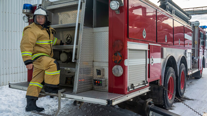 A firefighter in special clothes stands on the running board of a fire truck. A fire truck in winter. The rescue service. Fire truck and firefighter in protective clothing and helmet