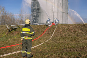Firefighters in heat-reflecting suits use a fire barrel to cool a burning tank of petroleum products at a petrochemical plant. Cooling of the tank with oil products after extinguishing the fire.