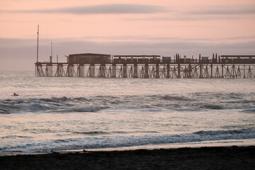 Surfer paddling out in the ocean alongside the long pimentel pier and beach, with a soft pastel sky at dusk