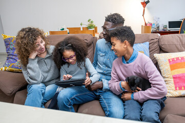 Happy multiracial family sitting on sofa, bonding over digital tablet with cheerful puppy, using technology together