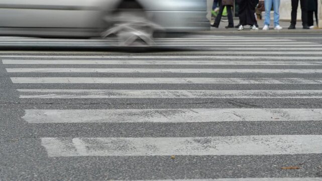 Low angle view of pedestrian zebra crossing on busy city street with cars driving by and people waiting on sidewalk. Urban traffic and transportation concept.