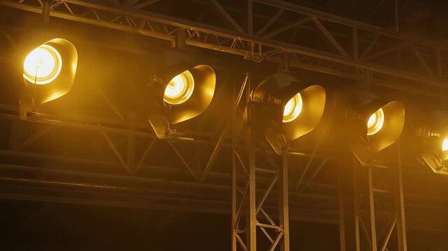 Row of bright stage lights mounted on a metal truss against a dark background with warm yellow lighting.