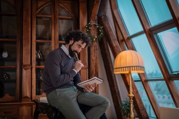 Man with beard thoughtfully writing in notebook in a cozy room with wooden beams and large window