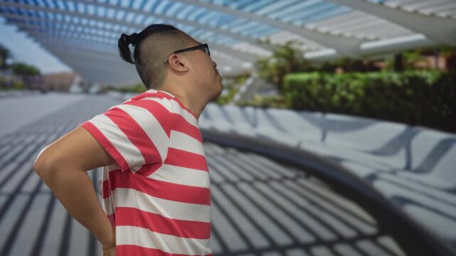 Young chinese man with glasses and manbun wearing red striped shirt leaning forward and holding lower back for backache on street under louvered canopy; discomfort.