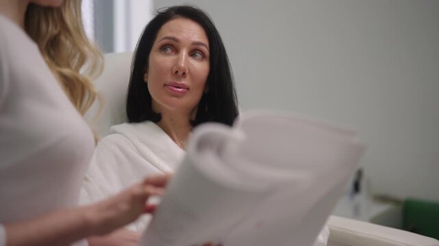 Cosmetologist consulting with female client in bathrobe at modern beauty clinic, reviewing documents and explaining personalized skin care and treatment options with professional care.