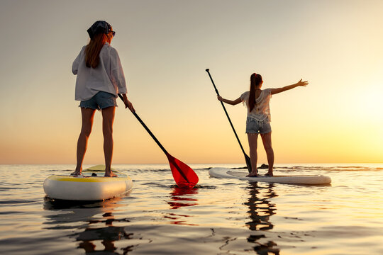 Two happy young women are standing on sup supsurfing boards at calm sunset lake