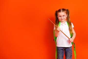 Young girl student with green backpack stands against bright orange background smiling while holding a stick in a playful school themed scene