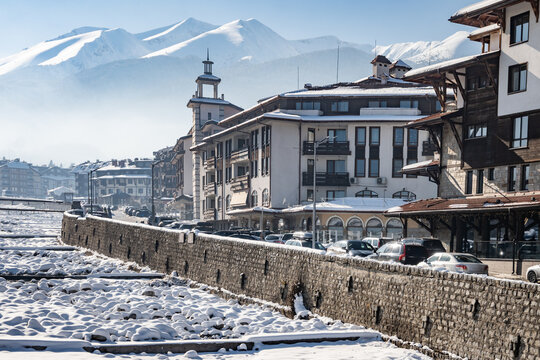 A panoramic look at a modern ski resort town in a snowy valley, featuring a stone embankment and buildings with the Pirin mountains in the hazy distance of Bansko, Bulgaria.
