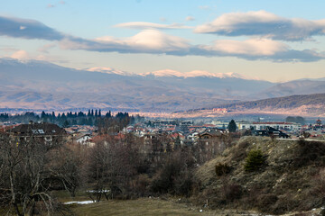 A sweeping panorama of Bansko town nestled in a valley, with the vast, snow-covered Pirin mountain range under a dramatic cloudy expanse.