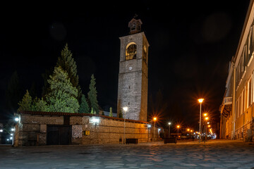 A stone bell tower is lit up at night, casting a warm glow on the surrounding cobblestone street and buildings in Bansko, Bulgaria.