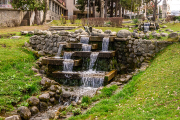 A charming tiered waterfall gently cascades over stacked stones and rocks, surrounded by lush green grass and trees, adding a natural water feature to an urban park in Razlog, Bulgaria.