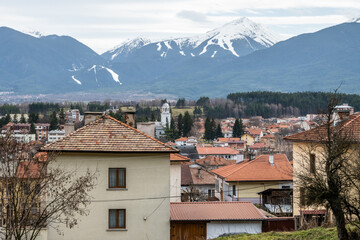An expansive urban landscape of Razlog featuring traditional red-tiled roofs and a prominent church tower, framed by distant snow-capped mountains and Bansko Ski Resort under a cloudy winter sky.