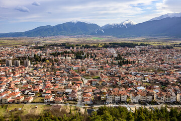 A sweeping, elevated panorama reveals Razlog town's detailed red-tiled rooftops, framed by distant snow-capped mountains and the sprawling green valley on a clear spring day.