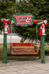 An outdoor wooden swing elaborately decorated with traditional red and white Martenitsa figures and green foliage, featuring a 'Chestitia Baba Marta' sign in Razlog.