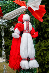 Detailed close-up of two handcrafted Martenitsa figures, one red and one white, made of yarn with intricate patterns, suspended outdoors against green pine foliage for Baba Marta in Razlog, Bulgaria