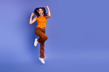 Joyful young female model jumping mid air in orange shirt and brown pants against blue studio background