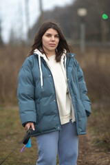 Young woman with long dark hair wearing a blue puffer jacket and gray sweatpants is walking a dog along a rural path surrounded by tall grass and trees in the background