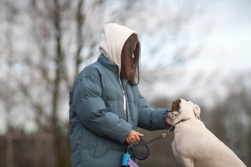 Young woman with long dark hair wearing a blue puffer jacket and gray sweatpants is walking a dog along a rural path surrounded by tall grass and trees in the background