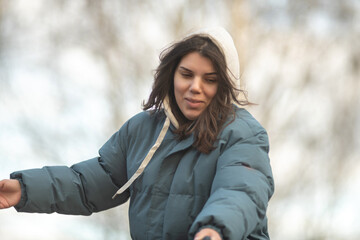 Young woman with long dark hair wearing a blue puffer jacket and gray sweatpants is walking a dog along a rural path surrounded by tall grass and trees in the background