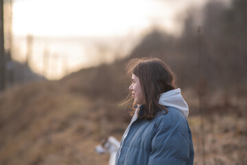 Young woman with long dark hair wearing a blue puffer jacket and gray sweatpants is walking a dog along a rural path surrounded by tall grass and trees in the background