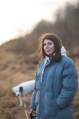 Young woman with long dark hair wearing a blue puffer jacket and gray sweatpants is walking a dog along a rural path surrounded by tall grass and trees in the background