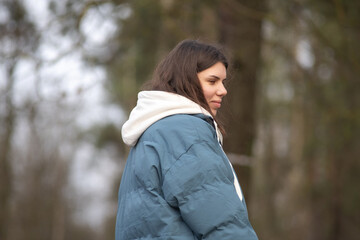 Young woman with long dark hair wearing a blue puffer jacket and gray sweatpants is walking a dog along a rural path surrounded by tall grass and trees in the background