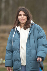 Young woman with long dark hair wearing a blue puffer jacket and gray sweatpants is walking a dog along a rural path surrounded by tall grass and trees in the background