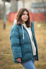 Young woman with long dark hair wearing a blue puffer jacket and gray sweatpants is walking a dog along a rural path surrounded by tall grass and trees in the background