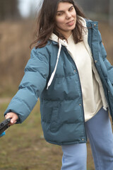 Young woman with long dark hair wearing a blue puffer jacket and gray sweatpants is walking a dog along a rural path surrounded by tall grass and trees in the background
