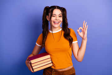 Young female student with book smiles and shows ok gesture against violet background