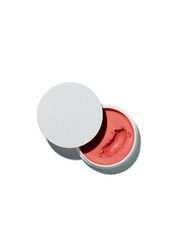Coral colored cream blush or tinted lip balm product sits open in a small white jar against a bright white background.
