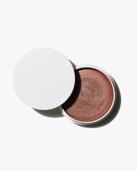 Open jar of shimmering bronze cream highlighter cosmetic product resting against a pure white background studio shot.