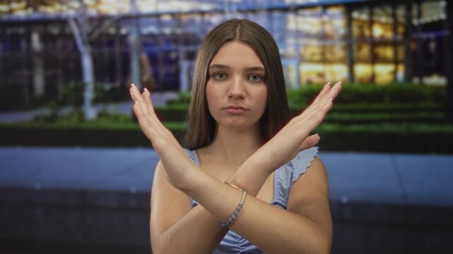 Teen girl crosses bare arms in x gesture under neon lights on a quiet evening city street at dusk; rejection.