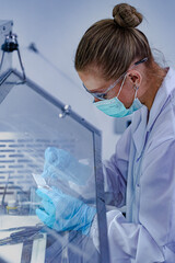 Close-up of focused female scientist wearing mask and safety glasses working in sterile fume hood. Researcher in blue gloves preparing tissue biopsy sample for histology analysis.