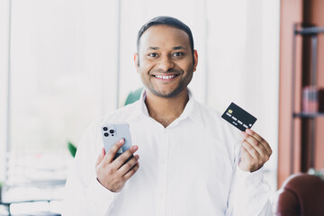 Young handsome business professional in bright office holds a smartphone and a credit card smiling at the camera in a modern loft interior setting
