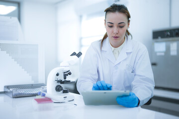 Caucasian female scientist using digital tablet to record data in modern laboratory. Researcher in white coat and blue gloves analyzing results near microscope.