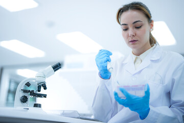 Caucasian female scientist using digital tablet to record data in modern laboratory. Researcher in white coat and blue gloves analyzing results near microscope.