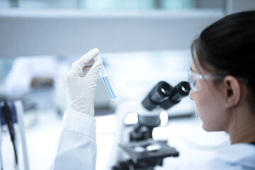 Female scientist holding test tube with clear liquid sample in modern lab. Researcher in white gloves inspecting chemical solution near microscope equipment.
