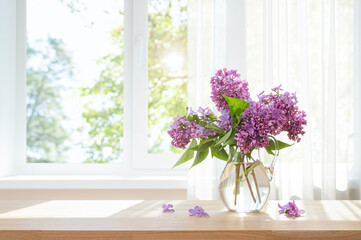 Bouquet of lilac flowers in glass vase on wooden table in front of summer day window and curtain background