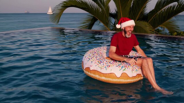 Happy man in santa hat floats on donut pool float. Christmas vacation in tropical pool. Man enjoys holiday swimming with inflatable donut. Summer christmas celebration at resort. Fun man laughs.