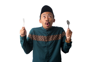 Excited Asian Muslim man wearing a green Indonesian baju koko and a black peci cap (songkok), pointing toward empty copy space while holding a spoon and fork in his hand, isolated on white background