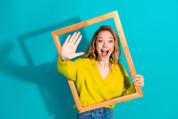 Young woman in yellow sweater waves through wooden frame against blue background smiling for...