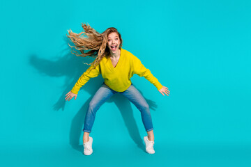 Young woman jumping with joy in bright yellow sweater and blue jeans against a blue background