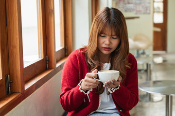 Happy Asian woman holding hot coffee cup with both hands in cafe. Smiling girl in red sweater enjoying aroma of fresh drink. Cozy winter lifestyle concept