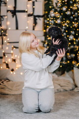 Young woman with blonde hair is joyfully holding a small black dog in a cozy indoor setting decorated with holiday lights and gifts, capturing a warm festive moment