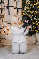 Young girl with long blonde hair kneeling on soft carpet, holding a small black dog in a cozy room decorated for the holidays with twinkling lights and a Christmas tree