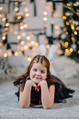 Young girl with brown hair, wearing a black dress, is lying on a soft carpet, smiling at the camera, surrounded by festive lights and holiday decorations, capturing a joyful moment