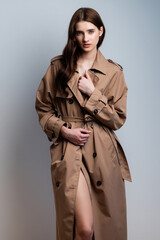 Elegant young fashion model wearing beige trench coat posing in studio on clean background, professional female portrait for modeling agency portfolio, modern commercial style photography