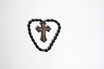 Black rosary beads arranged in a heart shape around a crucifix on a white background. Representation of faith, devotion, and religious love.